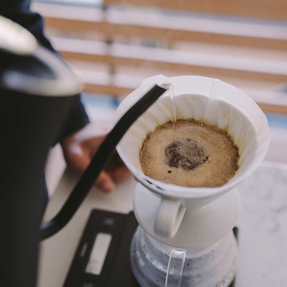 Ceramic white V60 brewing coffee and a black fellow kettle pouring water into the coffee bed