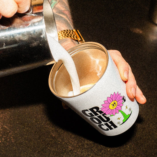 hands holding a stainless steel travel mug with espresso in it, pouring freshly steamed milk into the cup with a silver jug